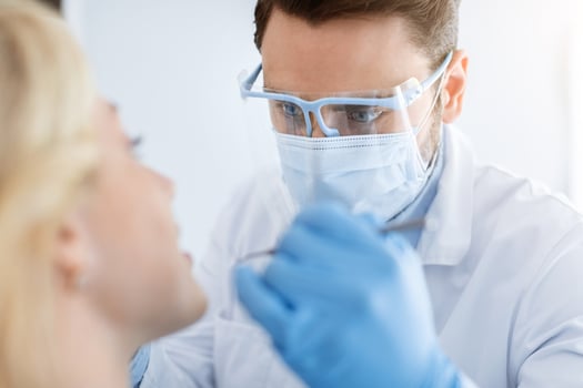 Closeup of young man dentist wearing medical face mask and protective glasses doing tratment or checkup for young blonde woman, wearing blue rubber gloves and holding dental tools photo