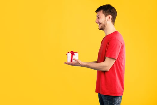 This Present Is For You. Side view portrait of happy young man holding wrapped gift box giving it to the side to copy space, isolated over blue studio background, greeting and celebrating special day photo