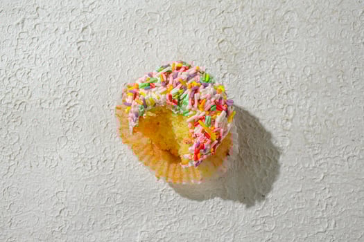 A colorful cupcake bitten topped with vanilla frosting and a generous amount of rainbow sprinkles, set against a textured white background for a clean and appealing look photo
