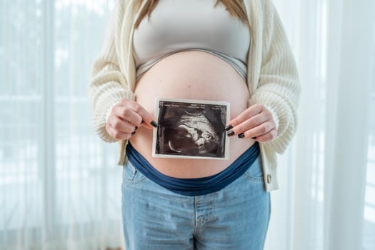 Young pregnant woman holding ultrasound scan photo of a fetus in front of her belly in third trimester at home. Concept of pregnancy, sonography. Maternity prenatal care