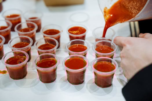 A person is diligently pouring sauce, likely a delicious condiment, into several plastic cups arranged neatly on a table, preparing for a culinary presentation or serving at an event photo