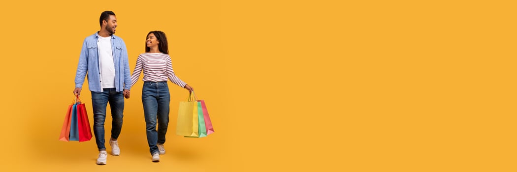 Smiling black couple carrying bright shopping bags, walking and holding hands against yellow studio background, happy young african american man and woman enjoying successful day of purchases photo