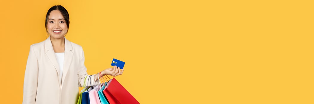 Smiling millennial chinese woman with shopping bags, credit card, enjoy sale, isolated on orange studio background. Finance for purchases, money recommendation for shopaholic photo