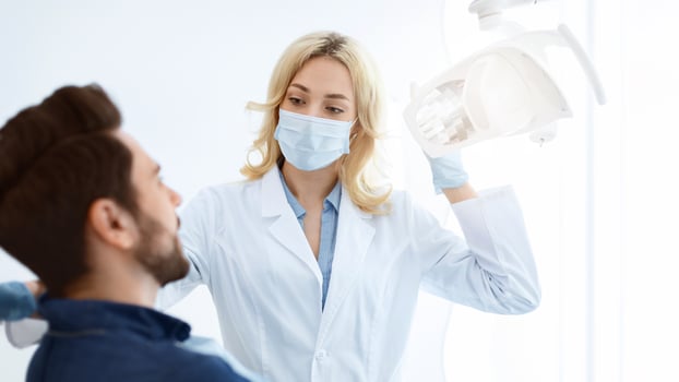 Female dentist blonde woman in face mask and medical gloves greeting male patient sitting in dental chair, turning on lamp while listening to bearded man complaints, closeup photo