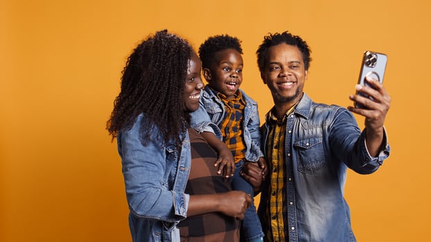 Sweet happy family with a small toddler take photos on smartphone, carrying their son and sharing embrace in studio. African american parents feeling grateful for their baby boy. Camera B.