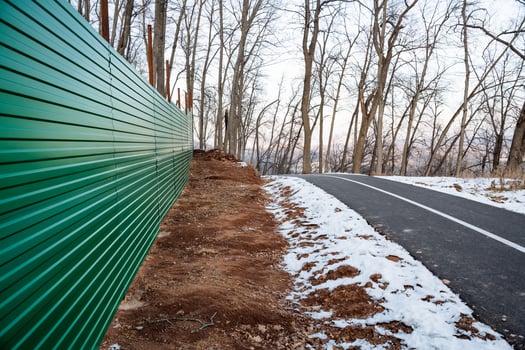 A vibrant green corrugated fence runs alongside a winter walking path set in a tranquil leafless forest, creating a picturesque scene for anyone enjoying the natural wilderness photo