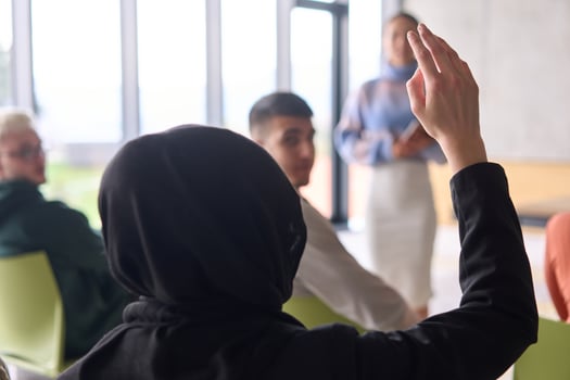 A Muslim businesswoman, dressed in professional attire and wearing a hijab, raises her hand during a business meeting to ask a question to the educator leading the session photo