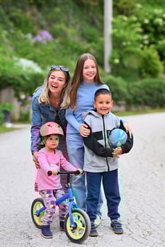 A happy group of people, including two women, a boy holding a ball, and a little girl on a balance bike with a helmet, enjoying outdoor activities on a paved path surrounded by greenery. photo