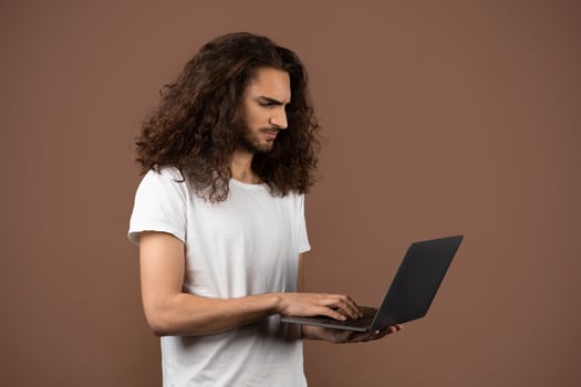 A young man with long curly hair stands alone, intently using a laptop. He is dressed casually in a white t-shirt, set against a warm brown background that enhances his concentration and lifestyle. photo