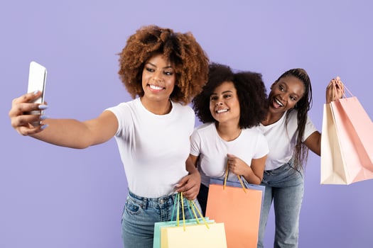 Three young African American women are smiling and posing for a selfie while holding colorful shopping bags. They showcase friendship and joy in a vibrant setting, celebrating their day out. photo