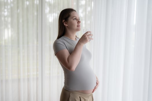 Pensive pregnant gen Z woman drinking water near window. Motherhood, pregnancy, hydration during pregnancy concept photo