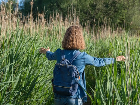 A view from the back of a blonde girl in a denim dress with a blue backpack on her back, walking along a path in the tall grass. Travelling in wild photo