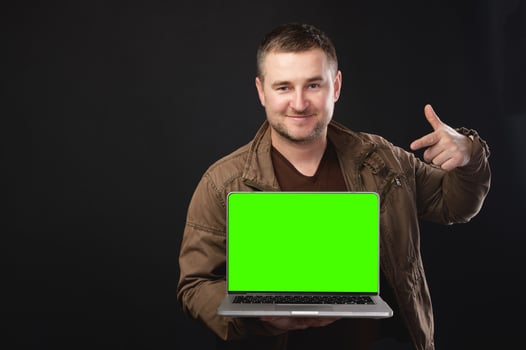 Young male IT specialist in leather jacket holding work on blank laptop screen isolated on plain green background. Smiling man gesturing at computer, place for your ad photo