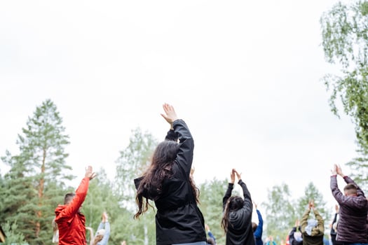 A joyful group of people gathers in a circle outdoors, engaging in a leisure activity under the sky surrounded by trees, nature, and green grass. They raise their arms in excitement photo