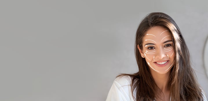 A woman is enjoying her skincare routine, smiling while applying products to her face. The setting is cozy, focusing on self-care and relaxation in a private space. photo
