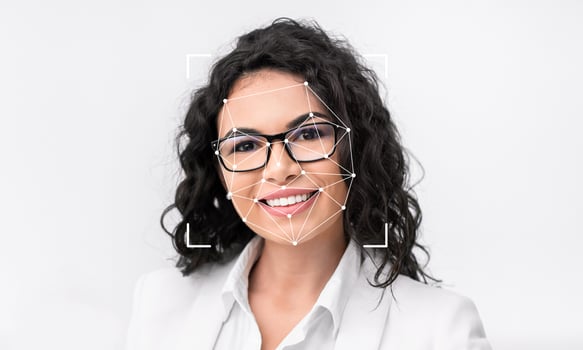 A smiling woman with curly hair and glasses stands confidently against a plain backdrop. Facial recognition technology is evident as geometric markers highlight her features. photo