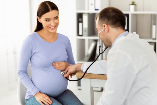 Gynecology Consultation. Smiling pregnant woman visiting her obstetrician doctor in maternity clinic, male gynecologist examining her belly with stethoscope, doing medical check-up photo