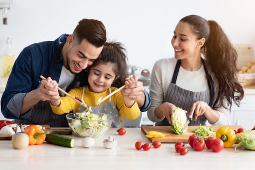 Portrait Of Cheerful Young Arab Family With Cute Little Daughter Cooking Together, Happy Middle Eastern Parents And Their Small Small Female Child Preparing Healthy Vegetable Salad At Home, Closeup photo