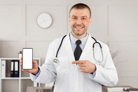Telemedicine Concept. Portrait of mature male medical practitioner holding smartphone with white blank screen for mockup template, pointing at gadget and smiling, wearing unifrom and stethoscope photo
