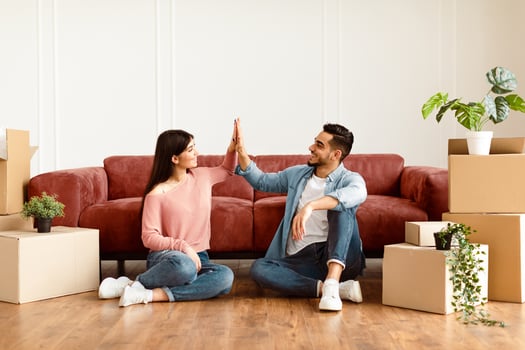 Good Job Concept. Happy young couple tenants giving high five, celebrating moving day, sitting on the floor near carboard boxes and plants. Smiling man and woman enjoying relocation photo