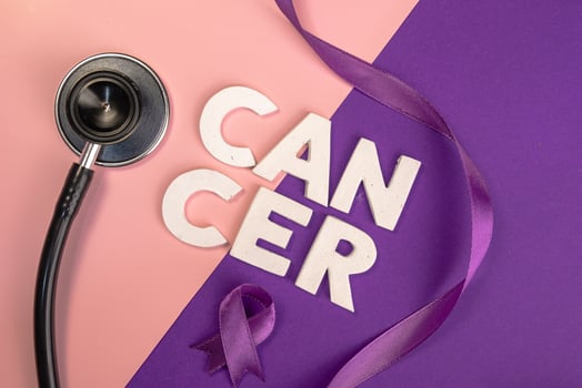 A stethoscope lies near the word CANCER spelled out in blocks next to a purple ribbon. World cancer day concept photo