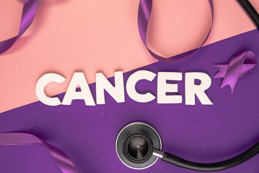 A stethoscope lies next to the word CANCER spelled out in white letters and a purple ribbon. International cancer day concept photo