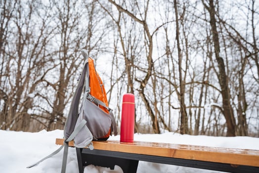 On a chilly winter day, a backpack and an insulated thermos are resting on a bench, surrounded by a blanket of soft, white snow, creating a picturesque and serene winter scene photo