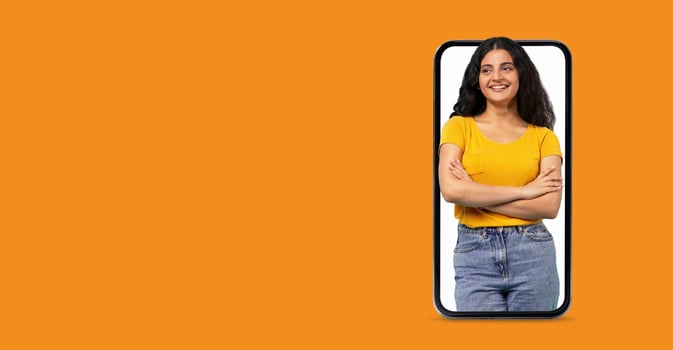 A young woman in a yellow shirt stands confidently with her arms crossed, showcasing a large smartphone screen. The bright background highlights her joyful expression while engaging with technology. photo
