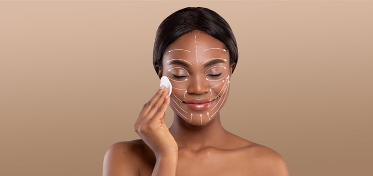 African American woman engages in a relaxing skin care routine, applying a product to her face with a gentle smile. She is isolated against a neutral background, highlighting her pampering moment. photo