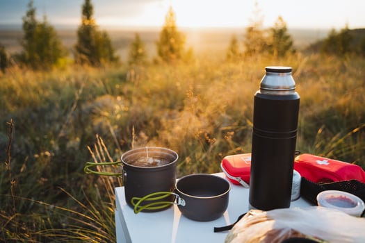 In a serene field, a rustic wooden table is beautifully set with pots, pans, and a warm thermos, inviting you to enjoy a meal amidst the natural beauty under the open sky photo