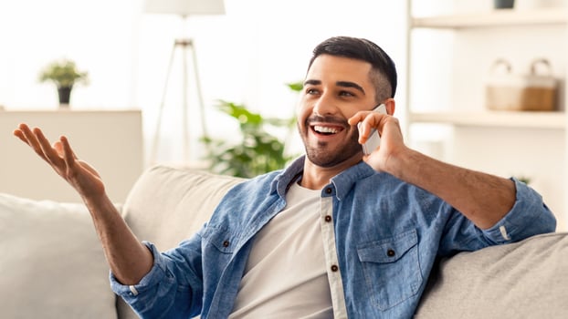 Distance Communication Concept. Portrait of excited smiling young israeli man talking with friends or family on mobile phone sitting on the couch, gesturing, selective focus, blurred background photo