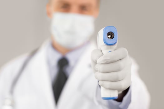 Concept of Covid-19 Prevention. Man in medical mask measuring body temperature. Doctor holding digital infrared non-contact thermometer in hand, pointing at camera. Coronavirus Symptoms photo