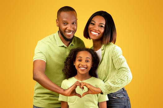 Happy loving family. Black parents holding hands in heart shape sign, posing with liitle daughter over yellow background and smiling together to camera photo