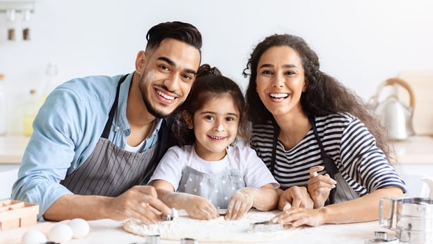 Cheerful arab father, mother and daughter making biscuits together, rolling pastry dough, middle-eastern mom and dad showing their little kid how to make cookies, wearing aprons, kitchen interior photo