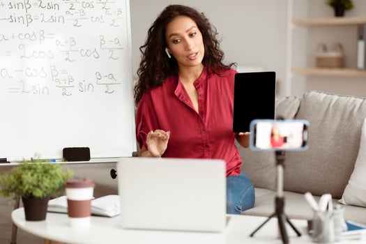 Lady math teacher making conference chat with students using laptop and cellphone on tripod, holding and showing tablet with empty screen with copy space for mockup, explaining photo