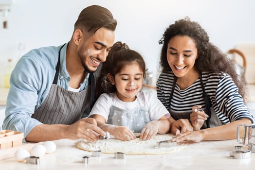 Positive arab father, mother and daughter making biscuits together, cutting pastry dough, middle-eastern mom and dad showing their little kid how to make cookies, wearing aprons, kitchen interior photo