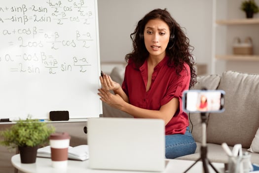 E-Learning. Confident woman having conference call using laptop. Lady teacher wearing wireless headphones talking to webcam, recording tutorial with mobile phone on tripod, streaming live photo