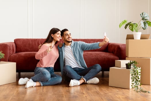 Happy young couple taking selfie in their new apartment, celebrating moving day, sitting on the floor near carboard boxes and plants. Smiling woman making showing v victory peace sign gesture photo