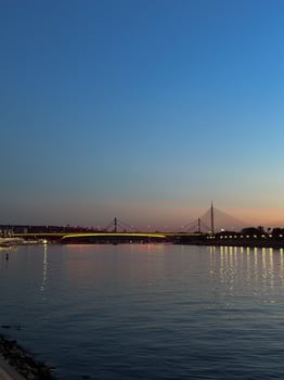 Twilight view of a lit bridge over the river in Belgrade, showcasing serene reflections on the calm water. Harmony of urban architecture in Serbia and nature in the evening fading. Danube river photo