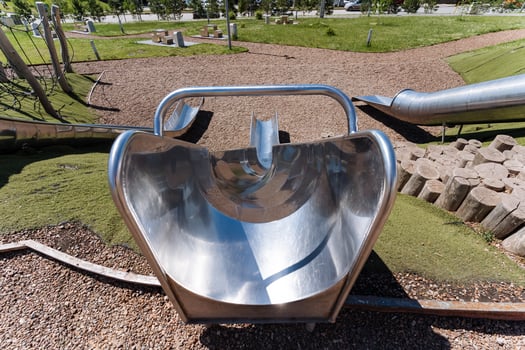 A stainless steel slide is featured at the playground in the park, surrounded by green grass and other elements like plants and trees. Safety equipment like helmets is encouraged for use photo