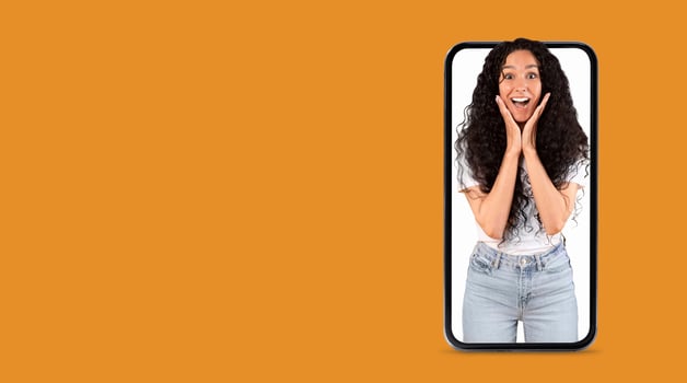 A person with curly hair joyfully reacts while appearing on a large smartphone screen. The vibrant orange background enhances the cheerful communication moment. photo