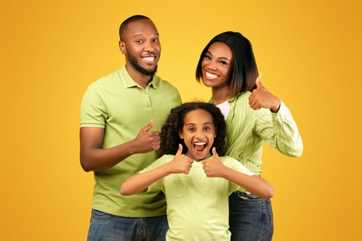 Best offer for families. Excited african american parents and their daughter showing thumbs up, recommending something, standing over yellow studio background photo
