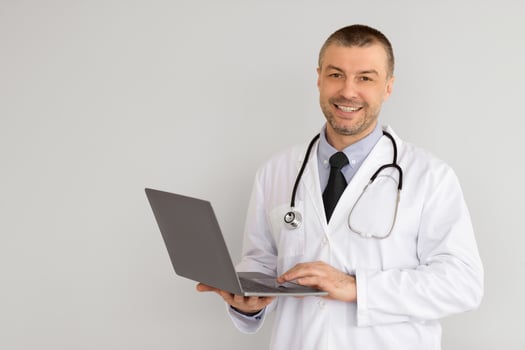 Health Care And Telemedicine Concept. Portrait Of Smiling Confident General Practitioner Wearing Coat And Stethoscope Holding And Using Laptop, Standing Isolated On White Studio Background, Copy Space photo