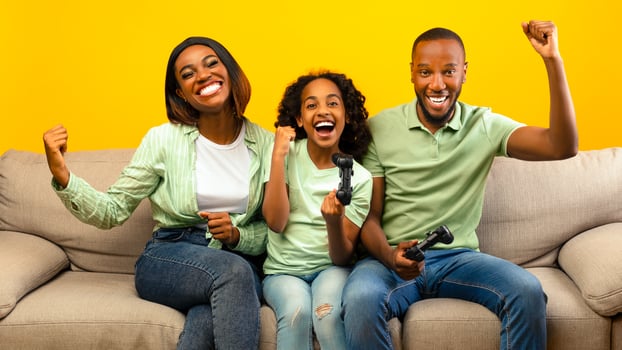 Family time activities. Happy black parents and their daughter playing games while sitting on sofa over yellow background. Dad, mother and child holding joysticks, winning game photo