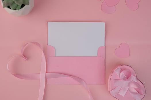 A pink envelope containing a blank card with a pink ribbon forming a heart shape next to a heart shaped gift box. invitation mockup design photo