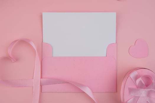 A pink envelope containing a blank white card next to a pink ribbon shaped as a heart and a pink gift box with a pink ribbon. invitation mockup photo