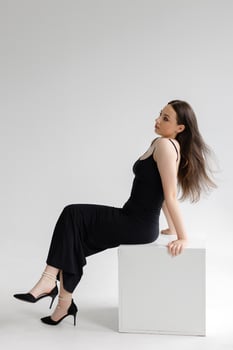 Fashion model posing sitting on white cube wearing black dress and high heels. photo