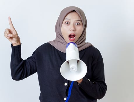 A surprised asian woman in a hijab passionately holds a megaphone while pointing upward photo