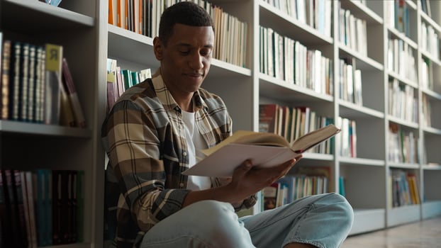 African American man sitting library floor bookcase reading book pleasure hobbies story knowledge learning student turning pages male guy academy study literary university history interesting reader photo
