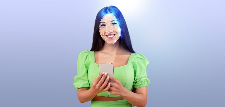 A young woman smiles while holding a smartphone, showcasing facial recognition technology. The background features an isolated, light color, highlighting her expression and the device. photo